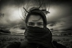[PHOTO: "Jessie," a CC-licensed image by LeTiger.] A young woman, nose and mouth covered with a scarf in foreground of flat, windswept landscape